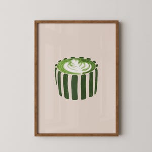 May include: A framed art print featuring a green matcha latte with latte art. The drink is in a striped cup, with a light beige background. The frame is a warm brown color.