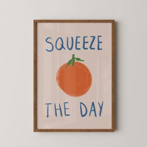 May include: A framed art print with the words "SQUEEZE THE DAY" in blue above and below an orange illustration of a citrus fruit with green leaves. The background is a light peach color, and the frame is brown.