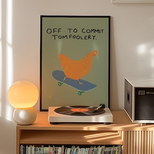May include: A framed poster with a chicken on a skateboard and the text "OFF TO COMMIT TOM FOOLERY" sits above a record player and a lamp. The poster has a green background and a black frame. The record player is white, and the lamp has a white base and a round, glowing bulb.