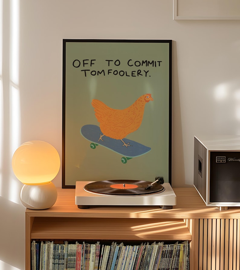 May include: A framed poster with a chicken on a skateboard and the text "OFF TO COMMIT TOM FOOLERY" sits above a record player and a lamp. The poster has a green background and a black frame. The record player is white, and the lamp has a white base and a round, glowing bulb.