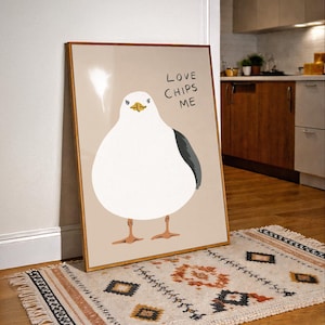 May include: A framed wall art print featuring a stylized illustration of a round white seagull with a dark gray wing and orange feet. The background is a solid light beige color. The text "LOVE CHIPS ME" is printed in black in the upper right corner.