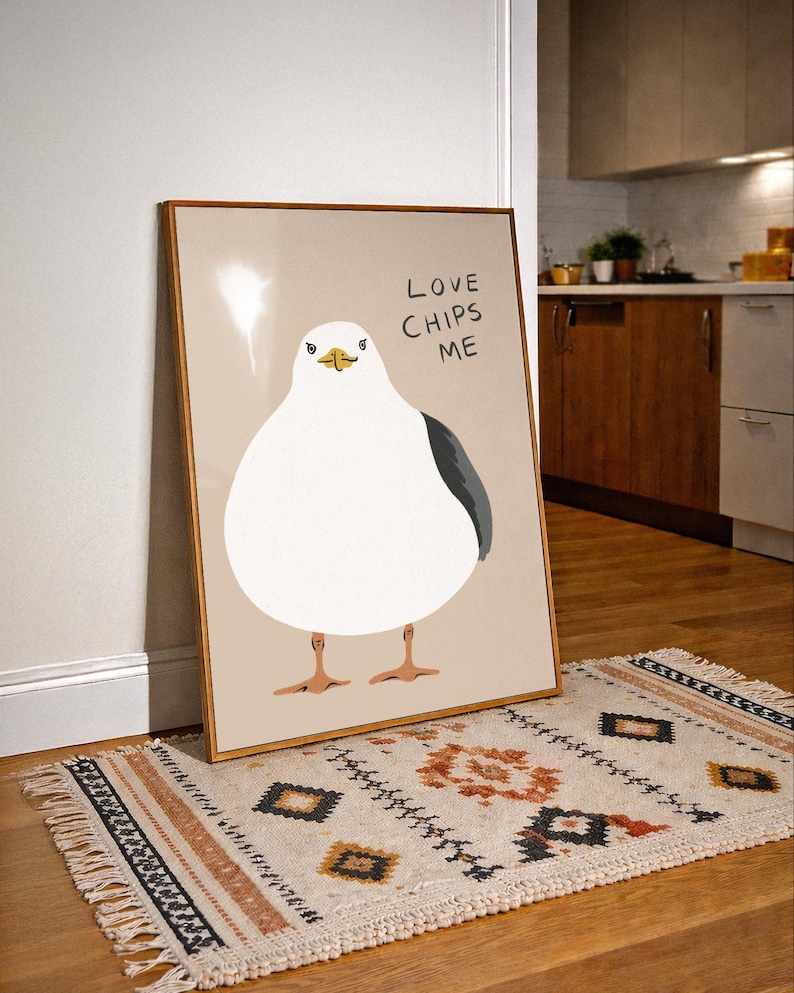 May include: A framed wall art print featuring a stylized illustration of a round white seagull with a dark gray wing and orange feet. The background is a solid light beige color. The text "LOVE CHIPS ME" is printed in black in the upper right corner.