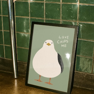 May include: A framed print featuring a white and gray seagull illustration on a sage green background. The text "LOVE CHIPS ME" is printed in white to the right of the bird. The seagull has orange feet and a yellow beak, presented in a black frame.
