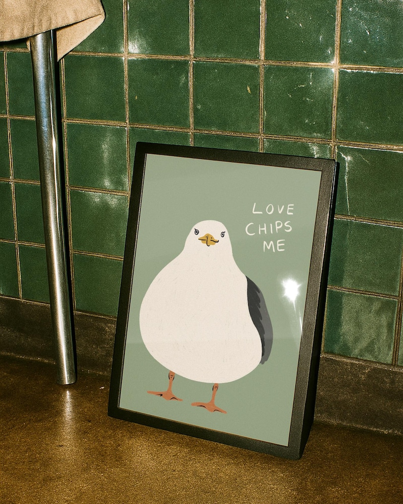May include: A framed print featuring a white and gray seagull illustration on a sage green background. The text "LOVE CHIPS ME" is printed in white to the right of the bird. The seagull has orange feet and a yellow beak, presented in a black frame.