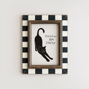 Könnte beinhalten: Ein gerahmter Druck mit einer schwarzen Katze, die sich dehnt, mit dem Text "Ooooooh Big Stretch". Der Rahmen hat einen braunen Innenrand und ein schwarz-weißes Schachbrettmuster am äußeren Rand. Das Kunstwerk wird auf weißem Hintergrund angezeigt.