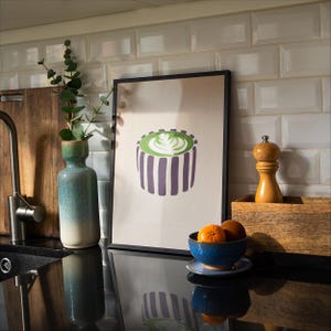 May include: A framed art print featuring a green and white latte design with purple stripes. The image is displayed on a kitchen counter with a blue and white vase, a wooden cutting board, and a bowl of oranges.