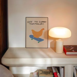 May include: A framed art print featuring a cartoon chicken on a skateboard, with the text "OFF TO COMMIT TOMFOOLERY." The chicken is orange, the skateboard is blue, and the wheels are green. The print is displayed on a white shelf with a lamp and books.