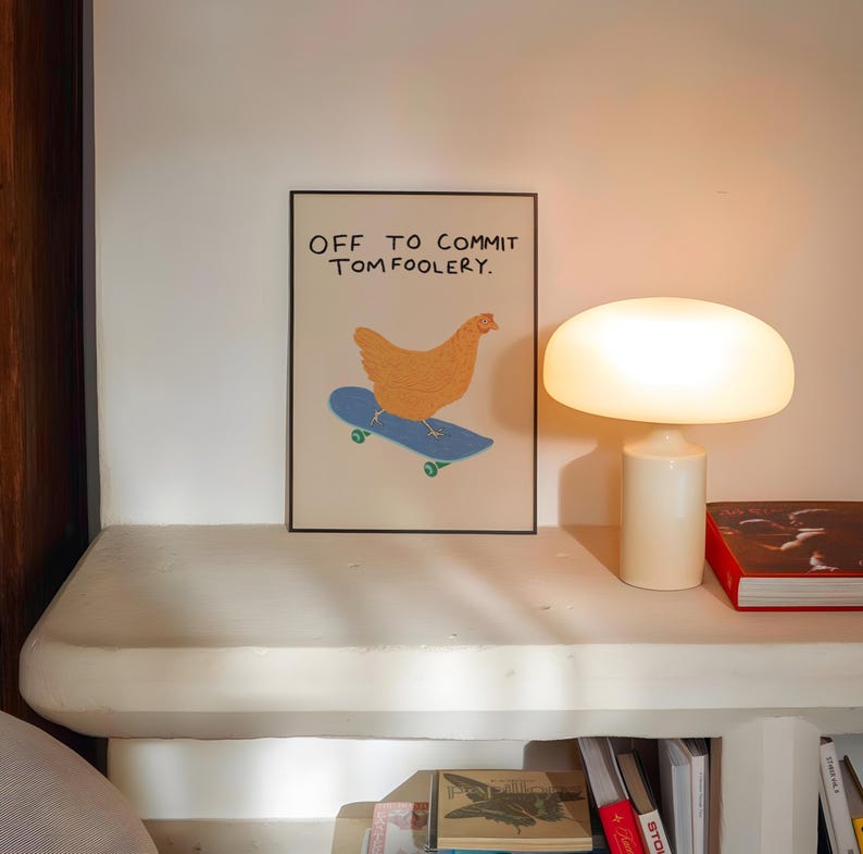 May include: A framed art print featuring a cartoon chicken on a skateboard, with the text "OFF TO COMMIT TOMFOOLERY." The chicken is orange, the skateboard is blue, and the wheels are green. The print is displayed on a white shelf with a lamp and books.