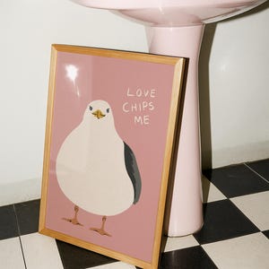 May include: A framed rectangular print featuring a stylized illustration of a white seagull with a dark gray wing on a dusty pink background. The text "LOVE CHIPS ME" is printed in white capital letters to the right of the bird's head.