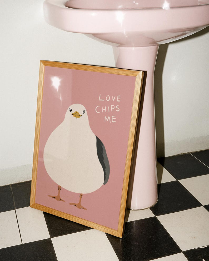 May include: A framed rectangular print featuring a stylized illustration of a white seagull with a dark gray wing on a dusty pink background. The text "LOVE CHIPS ME" is printed in white capital letters to the right of the bird's head.