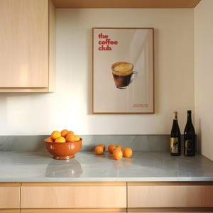 Puede incluir: Un mostrador de cocina con un taz&oacute;n de naranjas, dos botellas de vino y un p&oacute;ster enmarcado que dice "the coffee club". El p&oacute;ster presenta un dibujo de una taza de caf&eacute;.