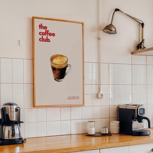 Puede incluir: Un p&oacute;ster enmarcado con un dise&ntilde;o minimalista que presenta una taza de caf&eacute; con espuma marr&oacute;n y blanca. El p&oacute;ster tiene el texto "the coffee club." y "fresh coffee every morning from 7am" en rojo.