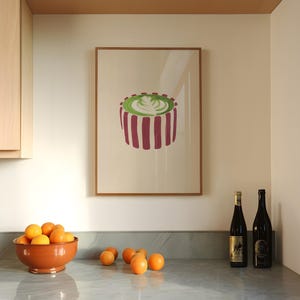 May include: A framed art print featuring a stylized image of a green and white latte with a floral design, set against a cream background. The print is framed in a light brown wooden frame. A terracotta bowl of oranges sits on a marble countertop.