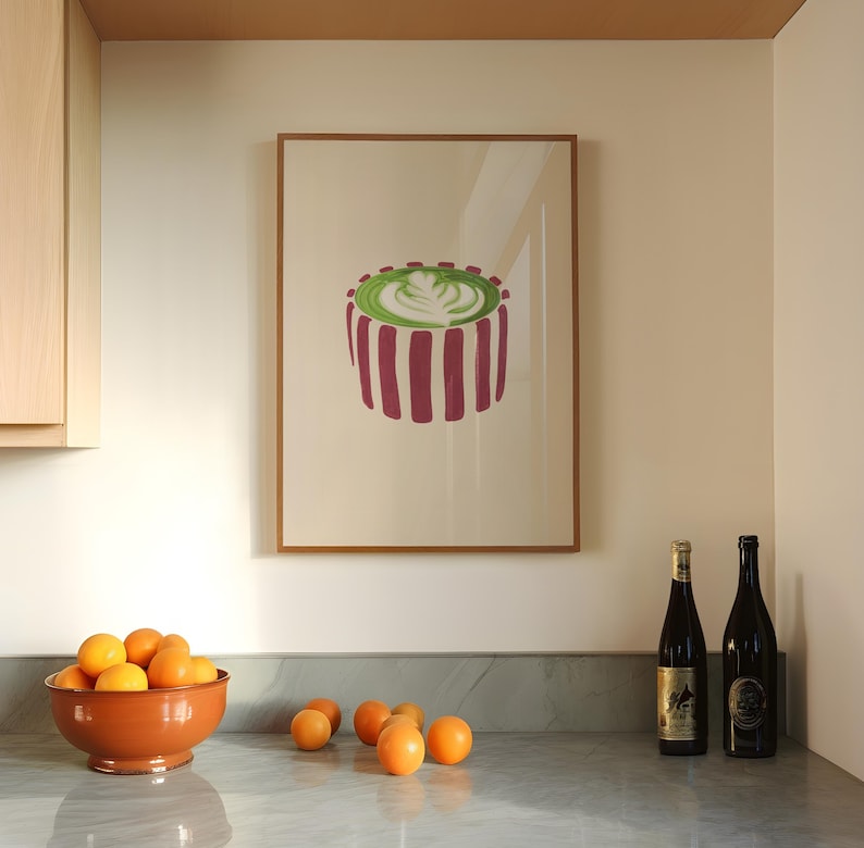 May include: A framed art print featuring a stylized image of a green and white latte with a floral design, set against a cream background. The print is framed in a light brown wooden frame. A terracotta bowl of oranges sits on a marble countertop.