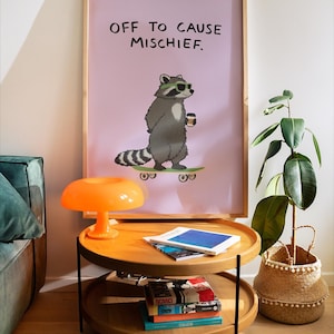 Puede incluir: Un estampado enmarcado con un mapache de dibujos animados con gafas de sol y una taza de caf&eacute; en un monopat&iacute;n, con el texto "OFF TO CAUSE MISCHIEF". El estampado est&aacute; encima de una mesa auxiliar de madera de dos niveles con libros y una l&aacute;mpara naranja.