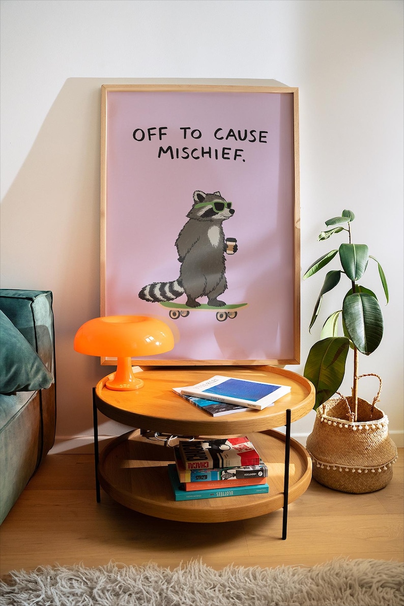 Puede incluir: Un estampado enmarcado con un mapache de dibujos animados con gafas de sol y una taza de caf&eacute; en un monopat&iacute;n, con el texto "OFF TO CAUSE MISCHIEF". El estampado est&aacute; encima de una mesa auxiliar de madera de dos niveles con libros y una l&aacute;mpara naranja.