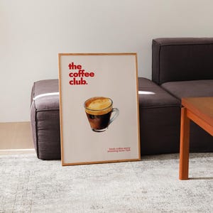 Puede incluir: Un p&oacute;ster enmarcado con el texto "the coffee club." en letras rojas. El p&oacute;ster presenta una ilustraci&oacute;n de acuarela de una taza de caf&eacute; sobre un fondo marr&oacute;n.