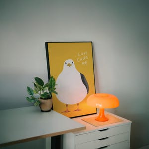 May include: A rectangular framed print featuring a white seagull with a grey wing on a yellow background, with the text LOVE CHIPS ME in white. Next to it is a small green plant in a tan pot and a bright orange mushroom-shaped table lamp.
