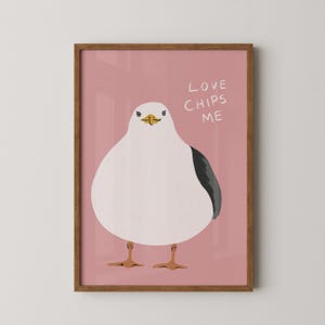 May include: A framed wall art print featuring a round white seagull with a yellow beak and dark gray wing on a dusty pink background. The text LOVE CHIPS ME is written in white capital letters to the right of the bird's head in this illustration.