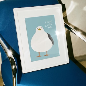 May include: A white framed print featuring a cartoon illustration of a round white seagull with a grey wing and orange feet against a light blue background. The text "LOVE CHIPS ME" is printed in white capital letters above the seagull's head.
