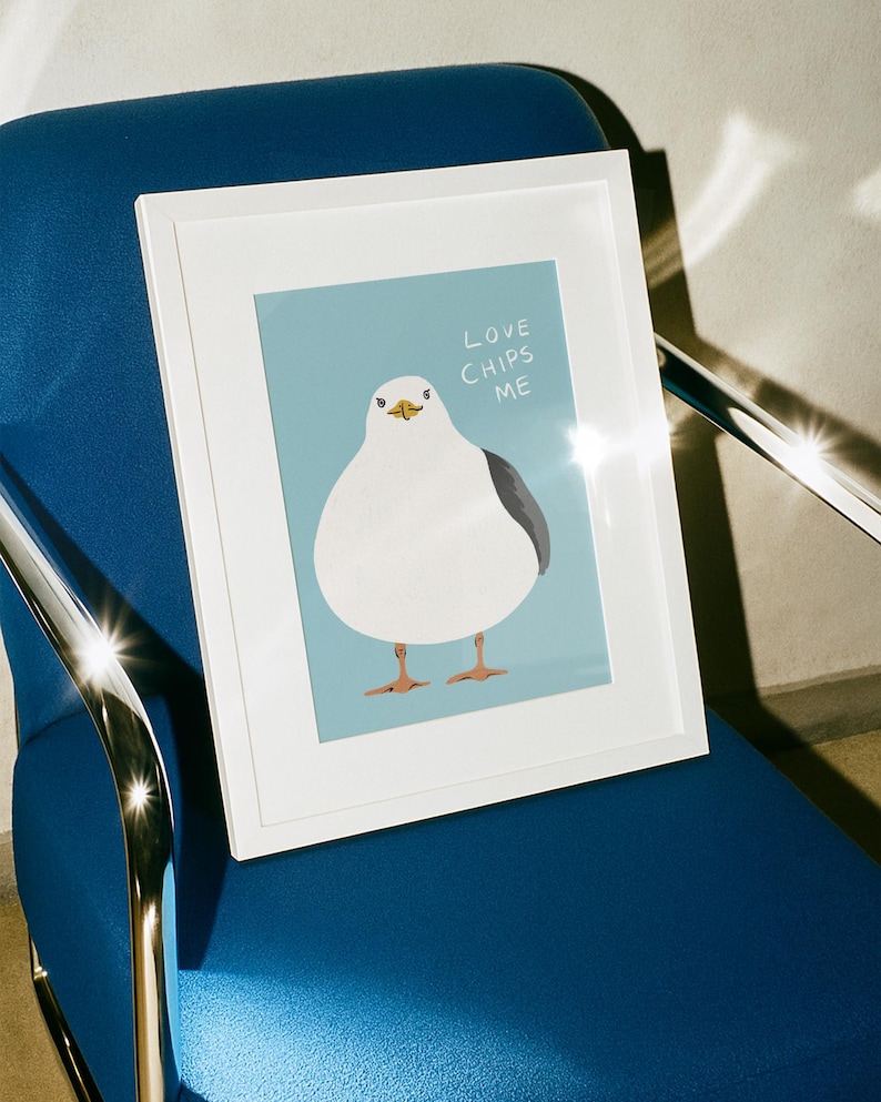 May include: A white framed print featuring a cartoon illustration of a round white seagull with a grey wing and orange feet against a light blue background. The text "LOVE CHIPS ME" is printed in white capital letters above the seagull's head.