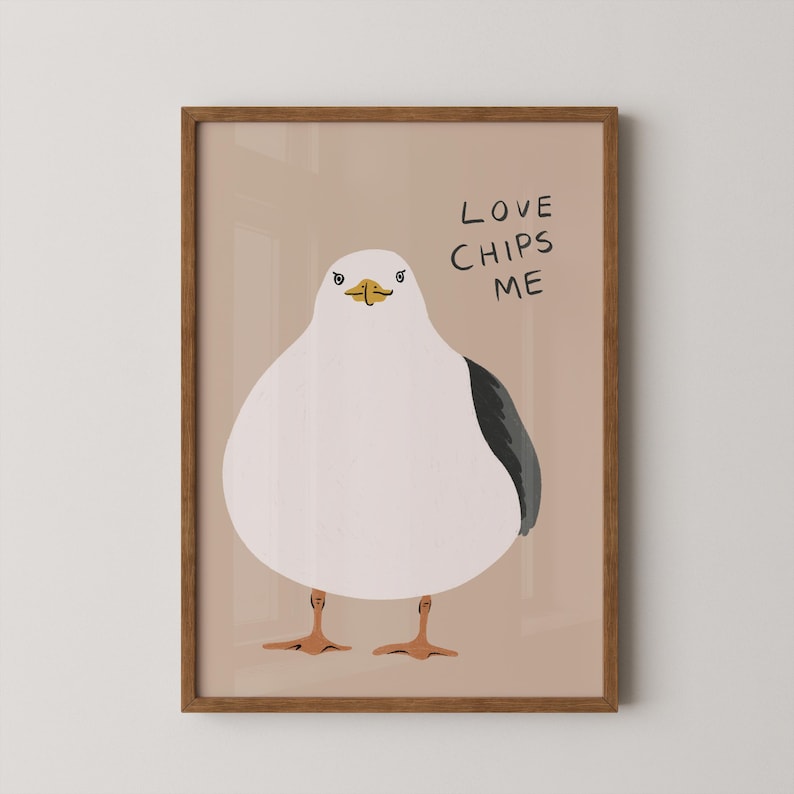 May include: A framed wall art print featuring a stylized illustration of a round white seagull with a dark gray wing and orange feet. The bird has a yellow beak and is positioned against a light tan background with the text LOVE CHIPS ME.