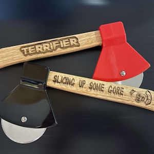 May include: Two wooden-handled pizza cutters shaped like axes. One has a red blade with the text "TERRIFIER" on the handle. The other has a black blade with the text "SLICING UP SOME GORE" on the handle.