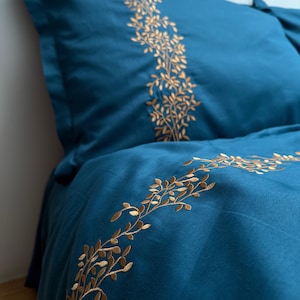 May include: Teal bedding set with gold embroidered leaf and branch patterns. The pillowcase and duvet cover display the botanical design, adding a touch of nature-inspired decor to the bedroom. The set is made of a soft material.