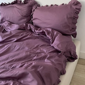 May include: A purple ruffled bedspread with matching ruffled pillowcases. The bedspread is made of a soft, smooth fabric and has a subtle sheen.