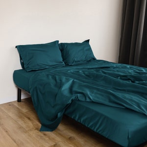 Peut inclure: Un lit bleu sarcelle avec un cadre en métal noir et deux oreillers assortis. Le lit est fait avec une housse de couette bleu sarcelle et se trouve sur un plancher de bois brun clair.