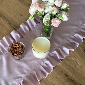 May include: A lilac satin table runner with a ruffled edge, adorned with a glass vase of pink and white flowers, a white candle with a gold rim, and a small glass bowl of almonds. The arrangement is set on a wooden surface.