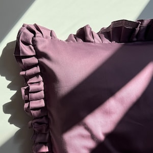 May include: A close-up of a mauve pillow with a ruffled edge. The pillow is made of a soft, smooth fabric and has a subtle sheen.