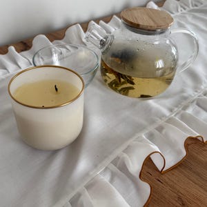 Op de afbeelding: Een witte kaars met een gouden rand, een glazen theepot met een houten deksel en een kleine glazen kom liggen op een witte tafel loper met ruches. De theepot bevat thee en kruiden.