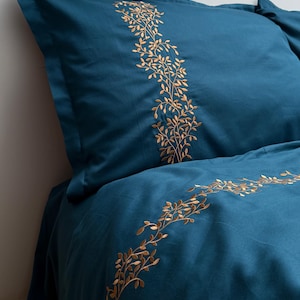 Könnte beinhalten: Türkisfarbenes Bettwäsche-Set mit goldfarbenen, gestickten Blatt- und Rankenmustern. Das Set umfasst einen Kissenbezug und einen Teil eines Bettbezugs, wobei die detaillierte Stickerei entlang der Kanten hervorgehoben wird. Der Stoff wirkt weich und luxuriös.