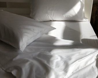 May include: A white bed with two white pillows and a white sheet. The bed is made and the pillows are fluffed. The sheets are smooth and wrinkle-free. The bed is in a room with a white wall and a wooden floor.