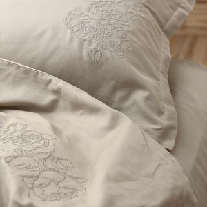 May include: A close-up of a beige bedding set with a floral embroidery design. The bedding is made of soft, comfortable fabric and features a delicate floral pattern.