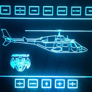 Op de afbeelding: Een blauw en wit digitaal display met een lijntekening van een helikopter en de tekst "AIRWOLF". Het display heeft knoppen voor verschillende functies, waaronder "FORE", "DEFENSIVE RADAR", "WEATHER RADAR", "GROUND RADAR", "TOPOGRAPHY", "ROTATE", "ATMOS", "WEAPON", "DEFENSE", "SLF DIG", "TEST", "DAY", "NIGHT", "VIDEO SCAN", "ZOOM-TELE", "ZOOM-WIDE" en "GRAPHICS-SUPER".