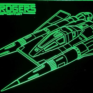 May include: A neon green light display featuring a line art illustration of a starfighter spaceship and the text Buck Rogers in the 25th Century against a solid black background. The design highlights the cockpit and dual engine nacelles.