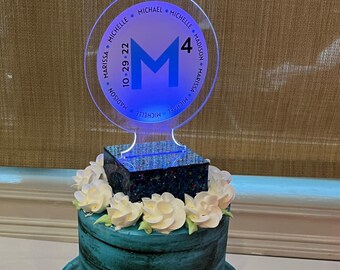 May include: A two-tiered blue and white cake decorated with white flowers. A clear acrylic topper with the letter "M" and the date "10-29-22" is on top of the cake. The topper also includes the names "Marissa", "Michael", and "Michelle" around the edge.