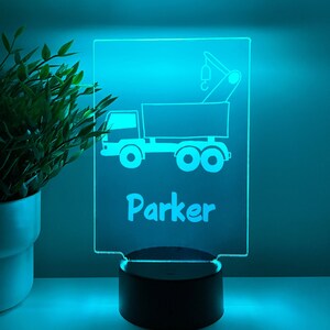 Könnte beinhalten: Ein blaues LED-Nachtlicht mit einer weißen Umrandung eines Kipplastwagens und dem Namen "Parker" darunter. Das Nachtlicht steht auf einem schwarzen Sockel.