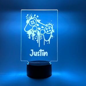 Könnte beinhalten: Ein weißes Acryl-Nachtlicht mit schwarzem Sockel. Das Licht zeigt eine weiße Silhouette eines Videospiel-Controllers mit Farbspritzern und dem Namen "Justin" darunter.
