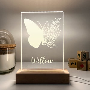 Può includere: Una luce notturna a LED rettangolare con un design a farfalla e floreale, e il nome "Willow" in corsivo bianco. La luce è posizionata su una base di legno ed è collegata a un cavo bianco. La luce è accesa, illuminando il design della farfalla.