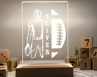 May include: A white silhouette of a football player holding a football with the name "OLIVER" next to him. The silhouette is on a clear acrylic panel with a wooden base. The panel is illuminated from behind.