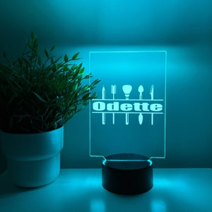 May include: A blue LED light with a clear acrylic panel featuring a white silhouette of makeup brushes and the name "Odette". The light is on a black base.