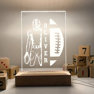 May include: A white silhouette of a football player holding a football with the name "OLIVER" next to him. The silhouette is on a clear acrylic panel with a wooden base. The panel is illuminated from behind.