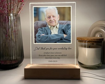 May include: A wooden base with a clear acrylic frame containing a black and white photo of a man. The frame has a white border and a light inside. The text "Dad, thank you for your everlasting love. A father's love is forever. Even after they pass, you can still feel their love every day. 1949-2019" is printed on the acrylic frame.