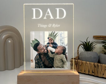 May include: A personalized LED photo frame with the word "DAD" illuminated in white. The frame features a family photo and the names "Thiago & Ryker" below the word "DAD". The base is made of light-colored wood.
