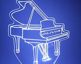 Può includere: Un contorno bianco di un pianoforte a coda con il nome "Olivia" sul lato, illuminato da una luce blu. Il pianoforte è su una base nera con un telecomando.