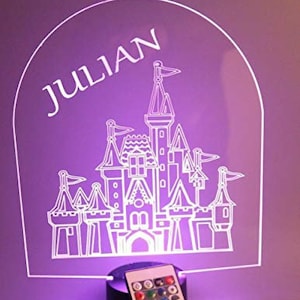 Puede incluir: Luz de noche LED púrpura con un diseño de castillo blanco y el nombre "JULIAN". Incluye un mando a distancia para cambiar los colores y una base negra.