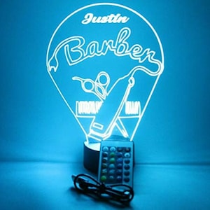 Puede incluir: Una luz nocturna LED blanca con un diseño personalizado que presenta la palabra "Barber" y el nombre "Justin". La luz tiene forma de lágrima e incluye un par de tijeras, un peine y una navaja. La luz está sobre una base negra y viene con un control remoto.